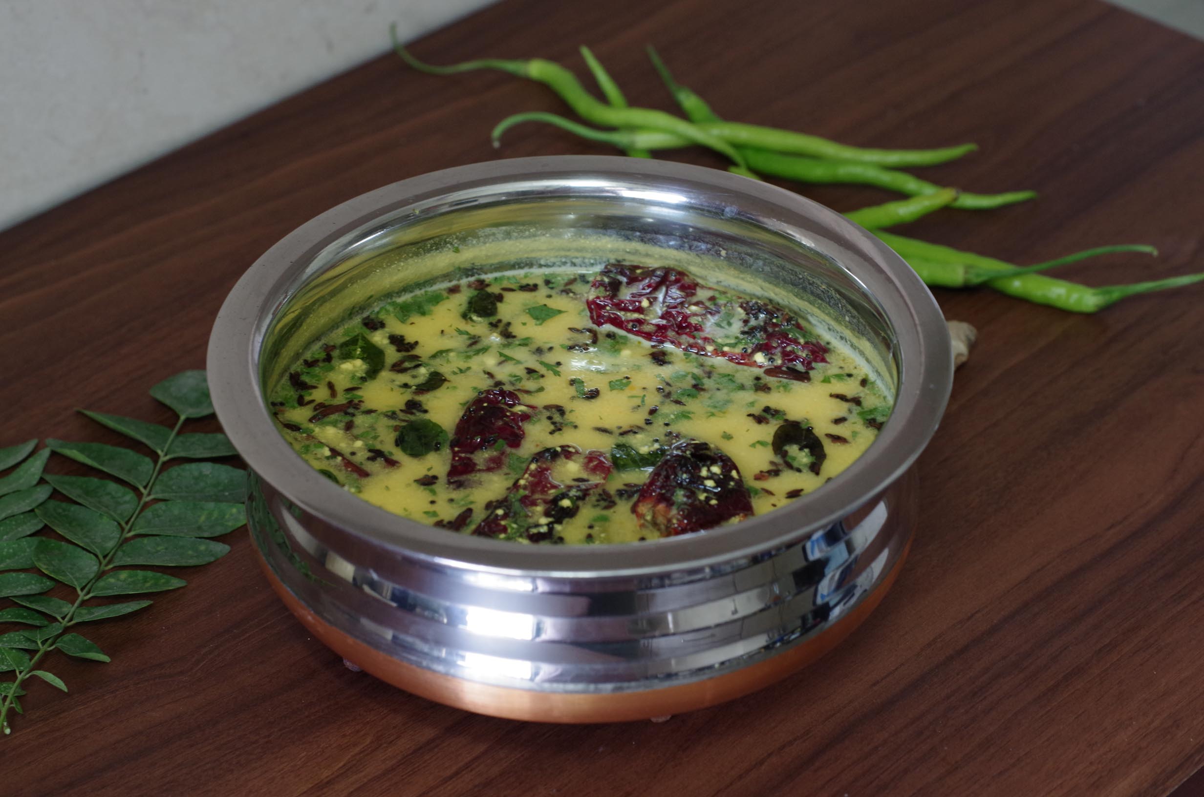 Gujarathi Kadhi in a bowl next to green chilies and curry leaves