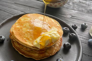 Flapjacks mit schmelzender Butter und Honig neben Blaubeeren auf einem schwarzen Teller - Flapjacks with melting butter and drizzled honey on a black plate with blueberries