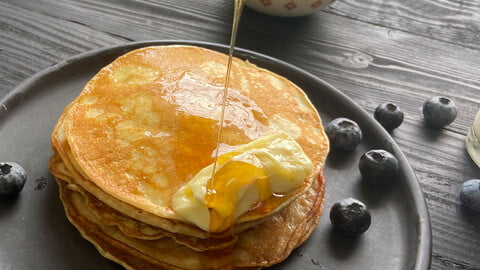 Flapjacks mit schmelzender Butter und Honig neben Blaubeeren auf einem schwarzen Teller - Flapjacks with melting butter and drizzled honey on a black plate with blueberries