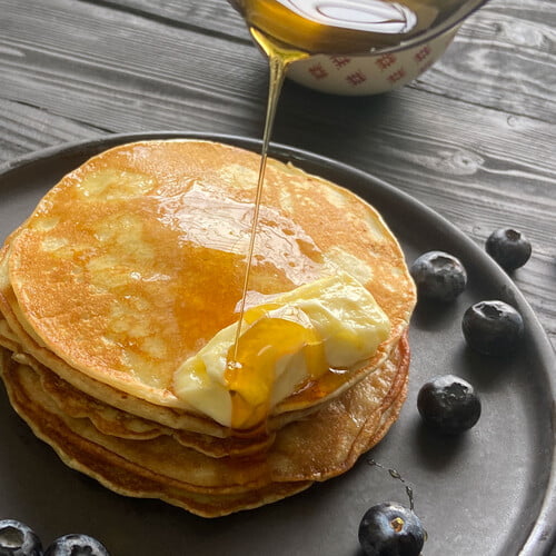 Flapjacks mit schmelzender Butter und Honig neben Blaubeeren auf einem schwarzen Teller - Flapjacks with melting butter and drizzled honey on a black plate with blueberries