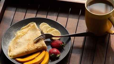 Crêpe mit Puderzucker und frischem Obst auf einem Teller angerichtet, neben einer Tasse Kaffee an einem sonnigen Nachmittag - Plated Crêpe with powdered sugar and fresh fruit next to a cup of coffee on a sunny afternoon