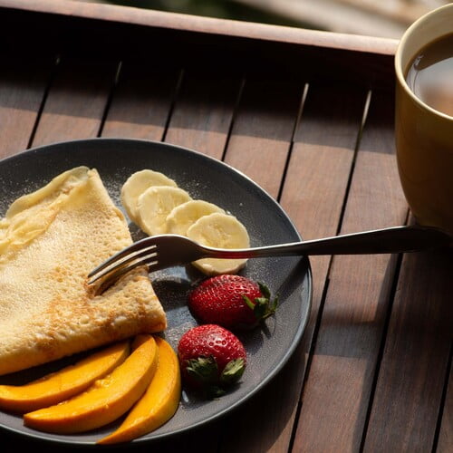 Crêpe mit Puderzucker und frischem Obst auf einem Teller angerichtet, neben einer Tasse Kaffee an einem sonnigen Nachmittag - Plated Crêpe with powdered sugar and fresh fruit next to a cup of coffee on a sunny afternoon