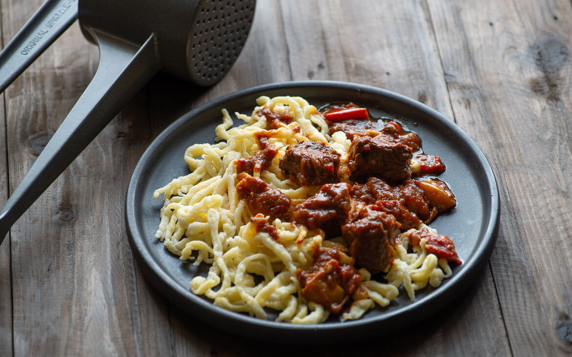 Spätzle mit Gulasch auf einem dunklen Teller neben meiner Spätzlepresse - Spaetzle with goulash on a dark plate next to my spaetzle press