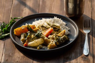 Avial auf einem Teller zusammen mit Reis. Neben dem Teller liegen ein Zweig Curryblätter und eine Gabel. Ein Trinkbecher aus Metall steht daneben. - Avial on a plate together with rice. Next to the plate is a sprig of curry leaves and a fork. A metal drinking cup stands next to it.