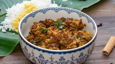 Beef Varutharacha Curry in einer verzierten Schüssel vor einem Bananenblatt mit Reis und Dal - Beef Varutharacha Curry in a decorated bowl in front of a banana leaf with rice and dal