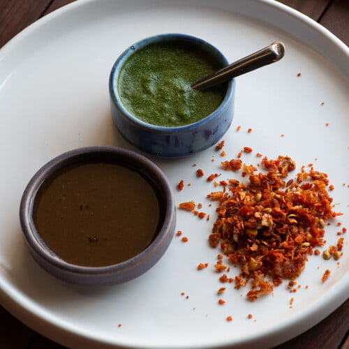 Indisches, trockenes Knoblauchchutney, grünes Chutney und süßes Chutney in kleinen Schalen auf einem weißen Teller - Indian dry garlic chutney, green chutney and sweet chutney in small bowls on a white plate