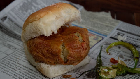 Batata Vada in a Pav with fried green chilies on the side on a newspaper