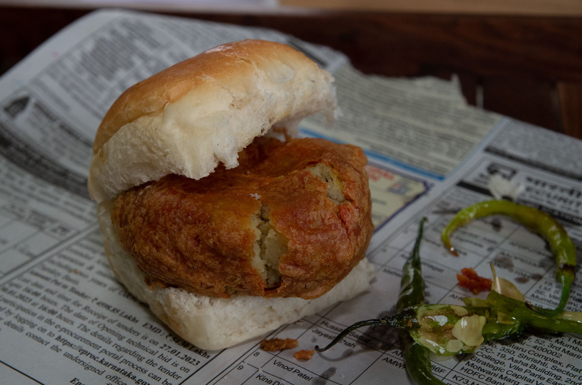 Batata Vada in a Pav with fried green chilies on the side on a newspaper