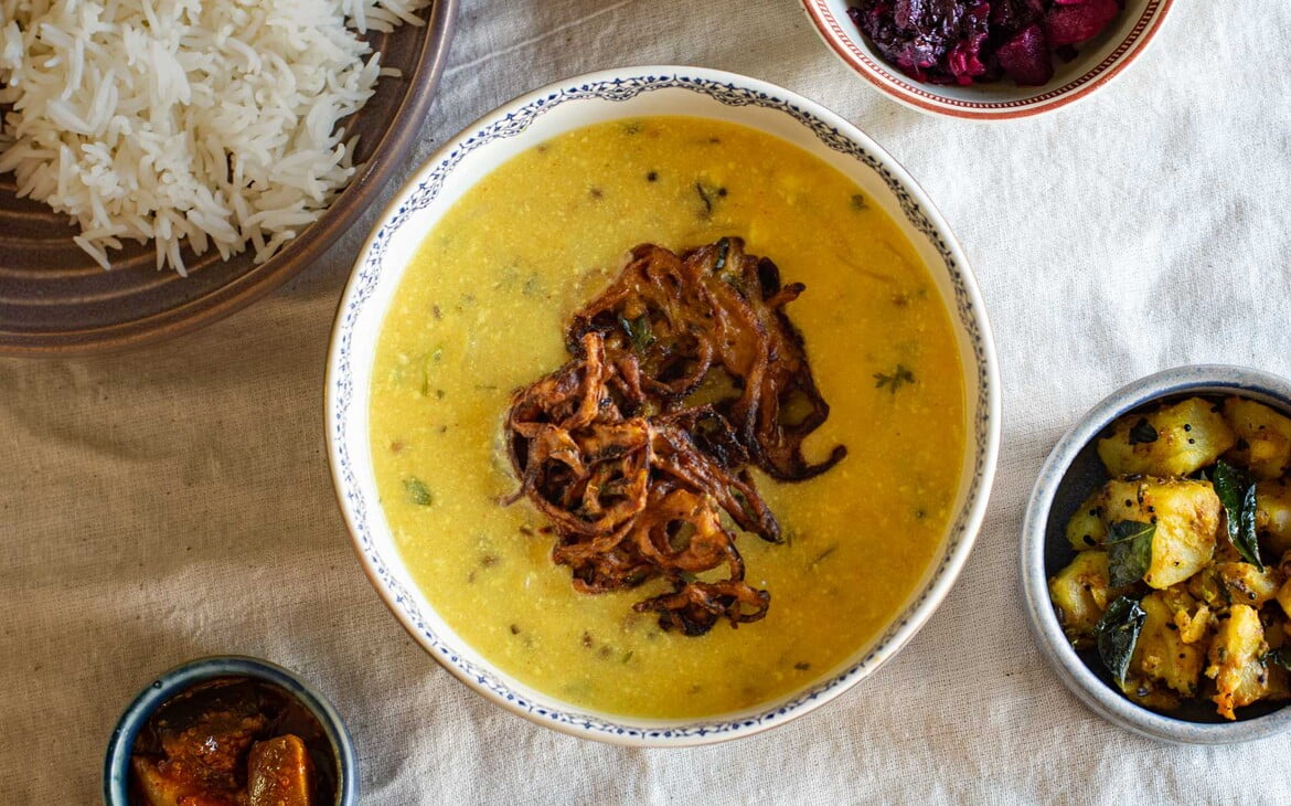 Kadhi mit Onion Pakora als Einlage in einer großen Schüssel. Um die Schüssel herum sind kleinere Schalen mit Mango Pickle, roter Beete, Kartoffel-Gemüse und einem Teller mit Reis - Kadhi served with Onion Pakora in a large bowl. Around the bowl are smaller bowls with mango pickle, beetroot, potato vegetable and a plate of rice