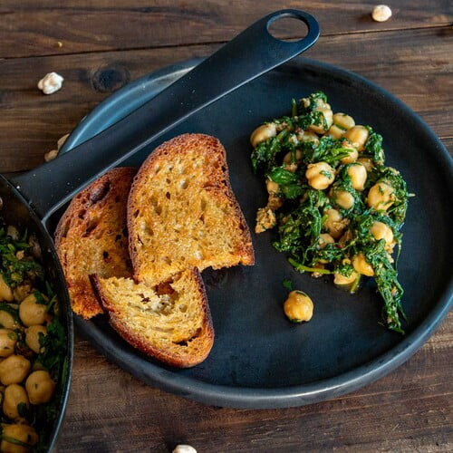 Espinacas con garbanzos zusammen mit in Olivenöl gebratenen Brotscheiben auf einem schwarzen Teller neben der Pfanne vom Kochen - Espinacas con garbanzos together with slices of bread fried in olive oil on a black plate next to the pan from cooking