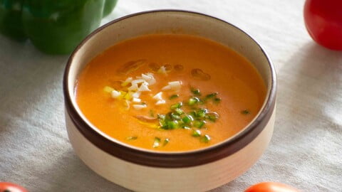 Eine Schüssel gekühlter Gazpacho auf einer weißen Tischdecke neben Tomaten und grüner Paprika - A bowl full of fresh, chilled Gazpacho on a white tablecloth next to ripe tomatoes and a green bell pepper