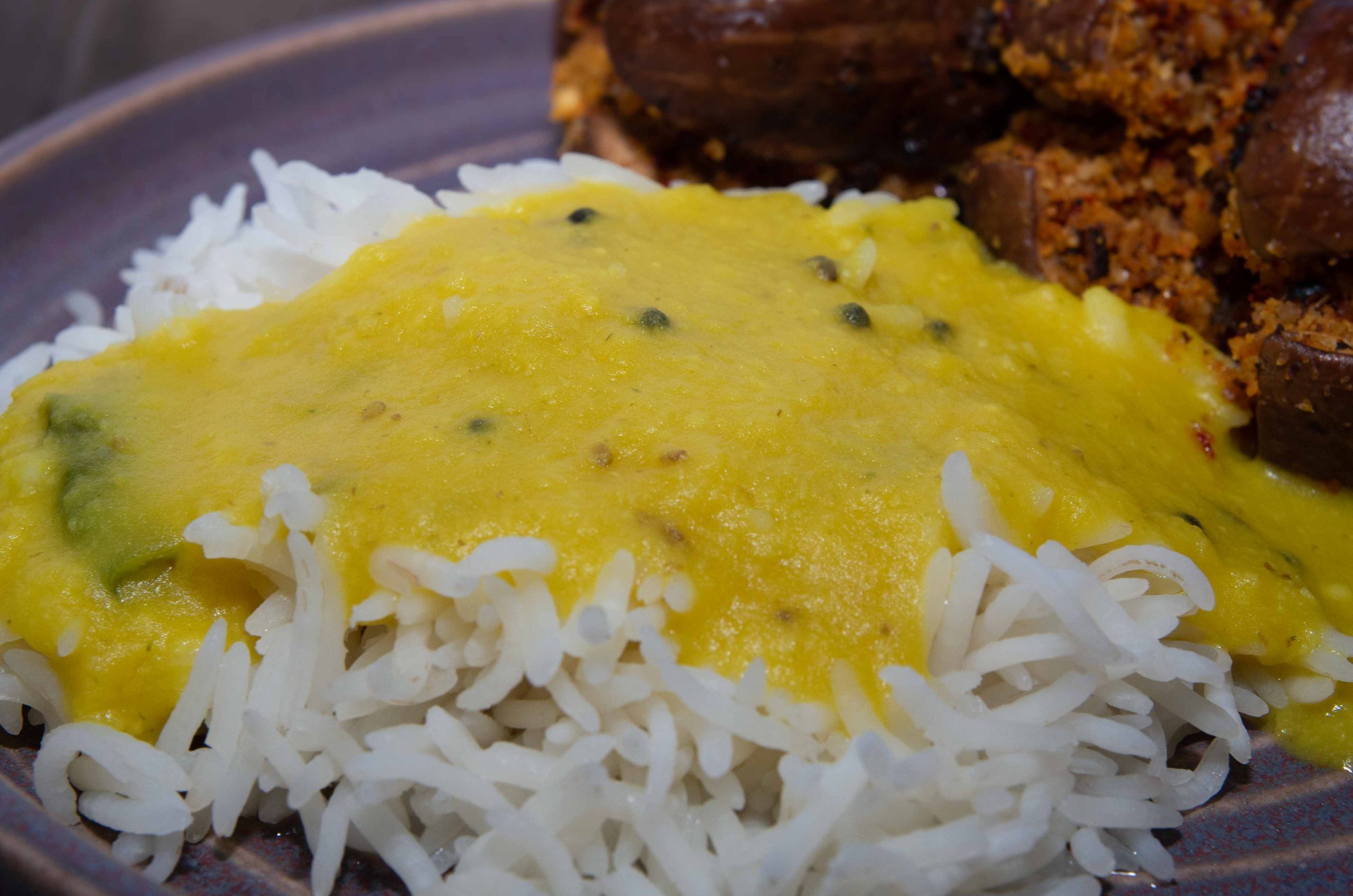 Vatpachi Dal - ein gelbes Toor Dal über eine Portion Reis gegossen, neben kleinen, mit Erdnüssen gefüllte Auberginen auf einem Teller angerichtet - Vatpachi Dal - a yellow toor dal poured over a portion of rice, arranged on a plate next to small eggplants stuffed with peanuts