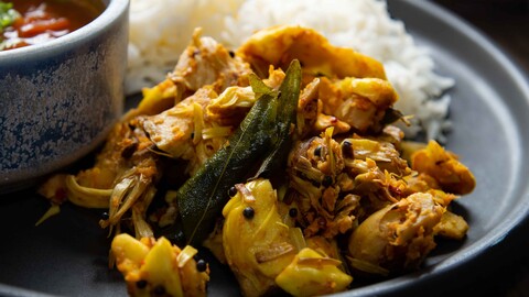 Südindisches Jackfruit Gemüse (Halasina Kayi Palya) auf einem dunklen Teller zusammen mit Reis und einer kleinen blauen Schüssel mit Tomato Rasam - South Indian Jackfruit vegetable (Halasina Kayi Palya) on a dark plate together with rice and a small, blue bowl of tomato rasam